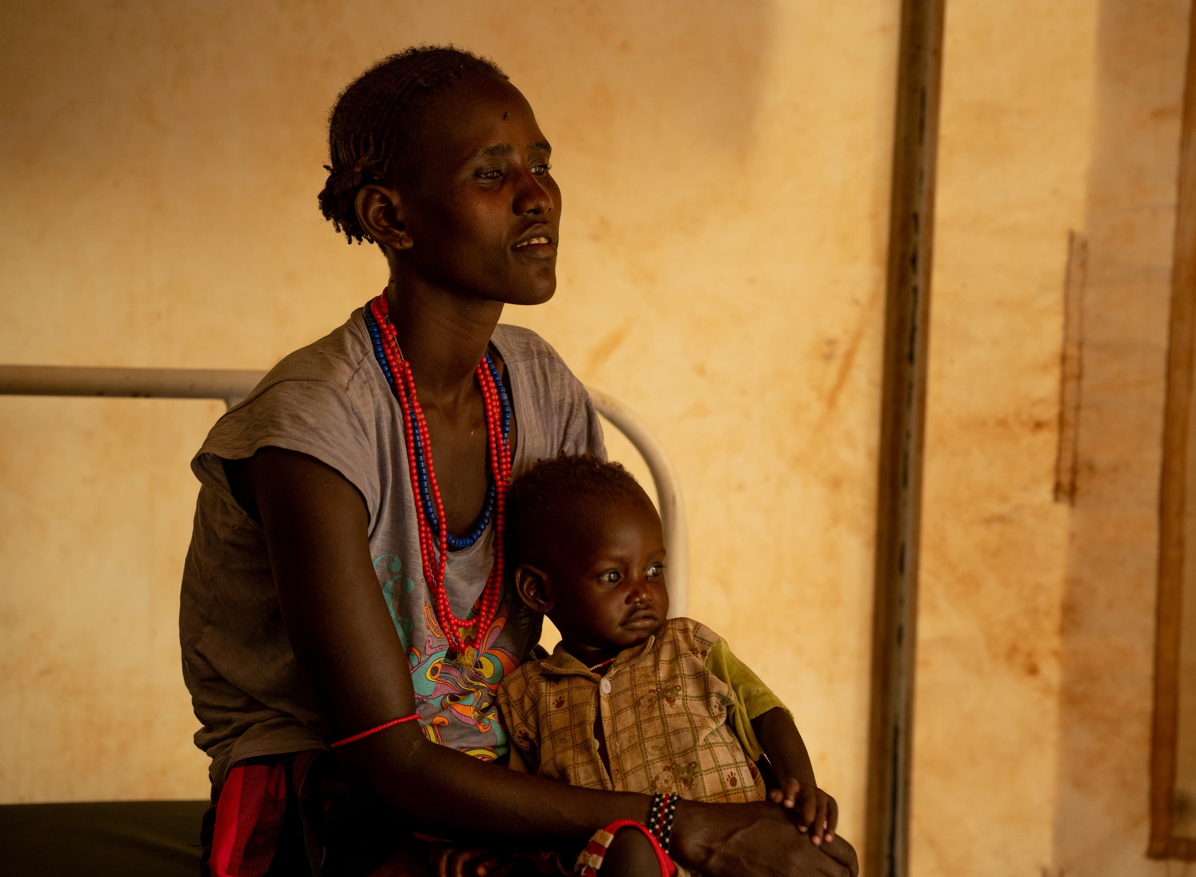 A woman holding a baby on her lap, seated on a bed inside a tent.