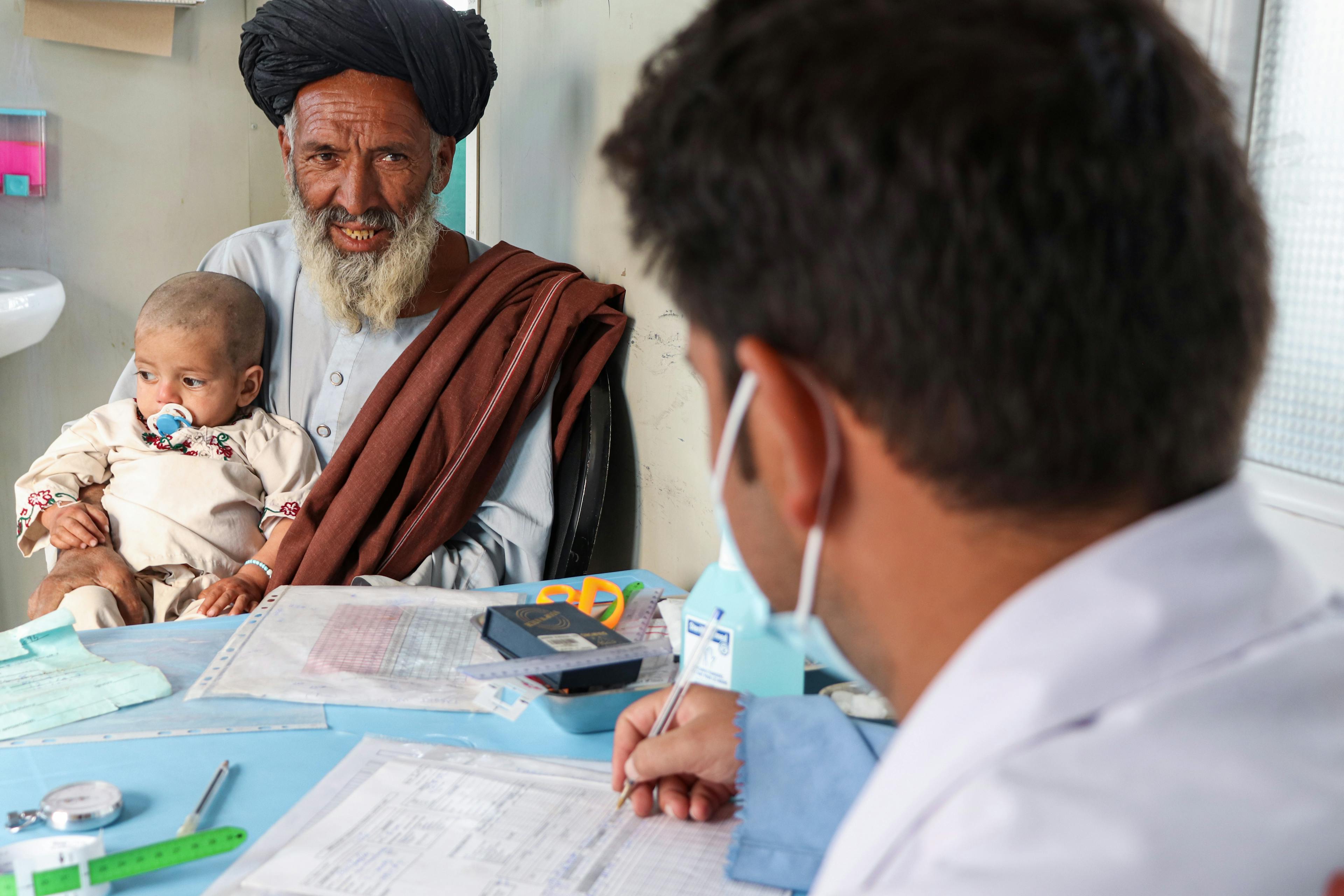 An old man holding a baby in his arms while talking to a doctor seen from the back. 